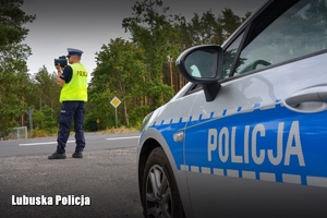 Napis policja na drzwiach radiowozu, w tle policjant kontroluje prędkość z jaką kierujący prowadzą pojazdy