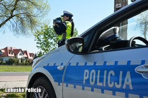 Napis policja na radiowozie, w tle policjantka kontroluje prędkość jadących pojazdów
