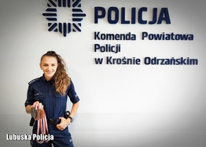 Policjantka w mundurze, trzymająca medale w dłoni. Na ścianie logo Policji i napis Komenda Powiatowa Policji w Krośnie Odrzańskim.