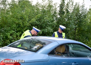 Jeden z policjantów rozmawia z kierowcą, drugi sprawdza dane pojazdu na mobilnym urządzeniu do sprawdzania danych w systemach policyjnych.