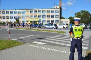 policjant przy przejściu dla pieszych z napisem Odłóż telefon i żyj , w tle osoby przy szkole