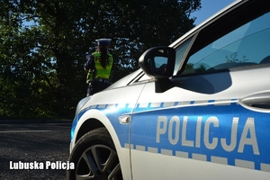 Napis Policja na drzwiach radiowozu, w tle policjantka kontroluje prędkość nadjeżdżających pojazdów