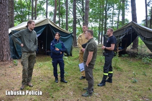 Przedstawiciele Policji, Straży Pożarnej i Nadleśnictwa Bytnica stojąc przy namiotach rozmawiają z mężczyzną.