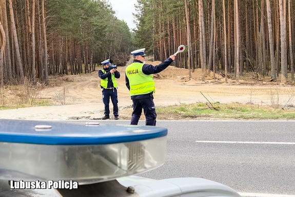 policjant na drodze zatrzymuje pojazd do kontroli, drugi z policjantów stoi przy drodze z urządzeniem